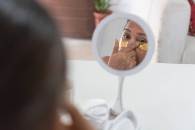 Olgun bir kadın evinde altın hidrojel göz bandı yapıştırıyor, küçük bir aynada kendine bakıyor.