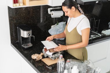 Ev hanımı, modern mutfağında geri dönüşüm için kağıt, plastik ve karton paketleme yapıyor.