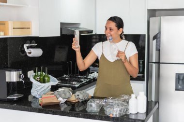 Latin bir kadın plastik şişe tutuyor ve telefon kullanıyor. Mutfaktaki geri dönüşüm ve atık ayrımı hakkında video çekiyor.