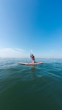 Güneşli bir günde ayakta kürek çeken bir adam, su sporu aktivitesinin tadını çıkarıyor.