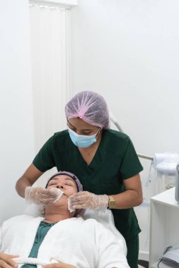 Güzellik uzmanı, güzellik kliniğinde yüz bakımı yaptıktan sonra cerrahi maske takıp kadın hastanın yüzünü temizliyor.