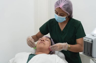Cerrahi maske ve eldiven takan Latin güzellik uzmanı güzellik salonunda hastanın yüzüne makyaj temizleyici pedleri uyguluyor.