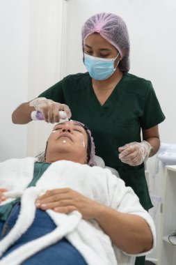 Cerrahi maske takan bir güzellik uzmanı ve bir hastanın yüzüne krem sürerek güzellik kliniğinde bir sedyede yatıyor.