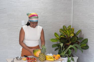 Latin kadın papaya soyuyor, ananas, muz, çilek ve portakallı tropikal meyve salatası hazırlıyor, renkli bir başörtüsü takıyor ve kulaklıkla müzik dinliyor.