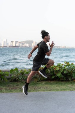 Genç atletik bir adam modern bir şehirde deniz kenarında koşuyor, sağlıklı yaşam tarzının tadını çıkarıyor.