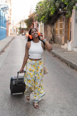 Turist bavulu çekiyor ve Cartagena de Indias 'ta yürürken kulaklıkla müzik dinliyor.