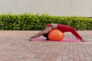 Genç bir kadın pilates egzersizi yapıyor. Açık havada spor salonunda spor topu kullanıyor.