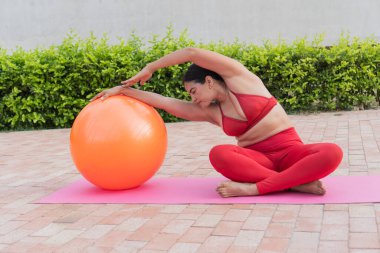 Pilates yapan kıvrımlı bir kadın, arka bahçedeki spor minderinin üzerinde spor topu ile yan esneme gösterisi, vücut pozitifliğini ve sağlıklı yaşam tarzını teşvik eden bir kadın.
