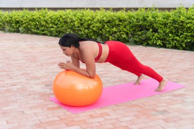 Genç Latin kadın pilates yapıyor. Açık havada bir paspasın üzerinde spor topu kullanıyor.