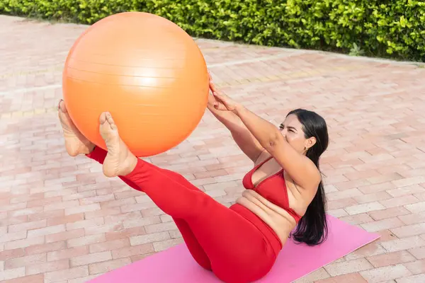 İspanyol kadın, açık havada pembe bir paspasın üzerinde turuncu bir fitness topuyla pilates yapıyor.