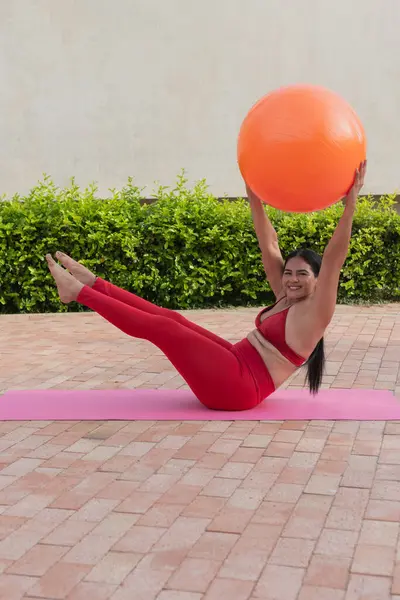 Kırmızı spor kıyafetli, kendine güvenen, iri yarı bir kadın arka bahçede yoga minderinde turuncu spor topuyla pilates yapıyor, vücut pozitifliğini ve sağlıklı yaşam tarzını destekliyor.