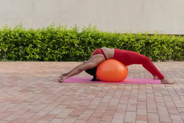 Genç bir kadın pilates egzersizi yapıyor. Açık havada spor salonunda spor topu kullanıyor.