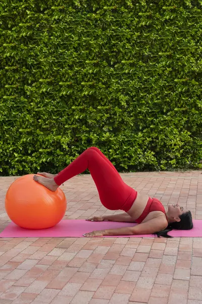 Beden eğitimi hocası, arka planda yeşil çit olan bir bahçede, pembe paspasın üzerinde turuncu İsviçre topuyla pilates yapıyor.