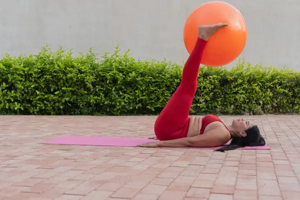 Kıvrımlı bir kadın spor topuyla pilates yapıyor, dışarıda paspas üzerinde egzersiz yapıyor.