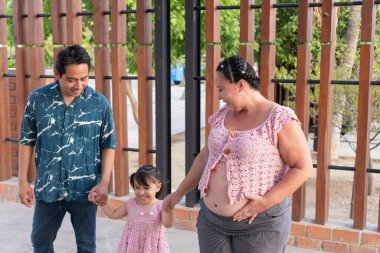 Mutlu İspanyol ailesi dışarıda eğleniyor. Hamile anne karnını ve kızının elini tutuyor.