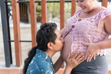 Hispanik bir adam hamile karısının karnını öpüyor. Bebekleri için şefkat ve beklentilerini dile getiriyor.