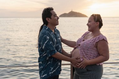 Mutlu İspanyol çift gün batımında deniz kenarında romantik bir anın tadını çıkarıyor, bebeklerini bekliyorlar.
