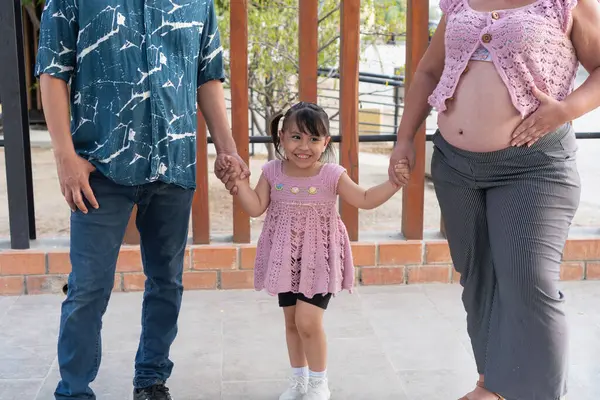 İspanyol ailesi dışarıda eğleniyor, hamile anne karnını tutuyor ve kız da ailesinin elini tutuyor.