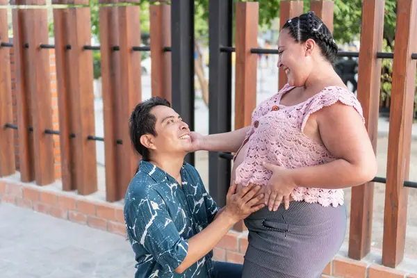 Mutlu İspanyol çift bebek bekliyor, müstakbel baba diz çöküp hamile eşinin karnına dokunuyor.