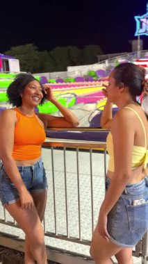 İki genç kadın, gece renkli bir lunapark gezisinin yanında gülüp sohbet ediyorlar.