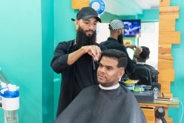 Kuaför erkek müşterinin saçını kesiyor sandalyede oturuyor, siyah pelerin giyiyor, güzellik salonunda.