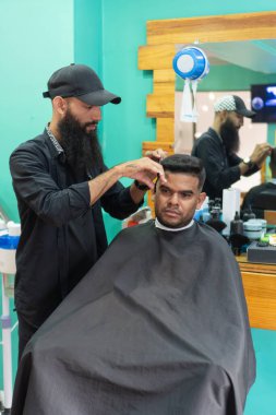 Kuaför berber dükkanında siyah pelerinli, sandalyede oturan erkek müşterinin saçını kesiyor.