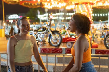 İki arkadaş lunaparkta eğleniyor, atlıkarıncanın yanında gülüyorlar.