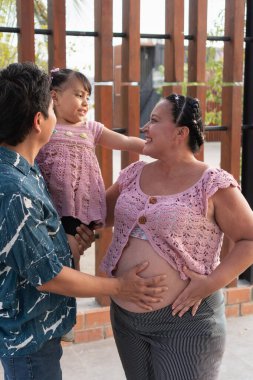 Mutlu İspanyol ailesi yeni bir bebek bekliyor, baba kızını kucağında tutuyor ve anne karnına dokunuyor.
