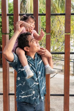 Genç İspanyol baba küçük kızını omuzlarında taşırken ikisi birlikte bir şeye bakıp işaret ediyorlar.