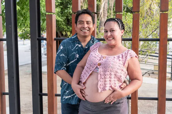 Mutlu İspanyol çift hamile karnını tutuyor, gülümsüyor ve parkta poz veriyorlar.