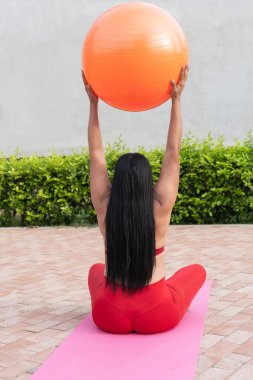 Kadın dışarıda pilates yapıyor, kolları havada turuncu bir fitness topunu kaldırıyor, pembe bir paspasın üzerinde oturuyor.