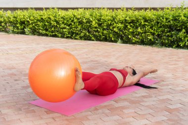 Genç bir kadın dışarıda bir paspasın üzerinde spor topu kullanarak pilates yapıyor.