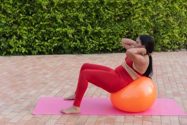 Büyük beden kadın vücut kaslarını güçlendiriyor, arka bahçede turuncu bir fitness topunda pilates yapıyor.