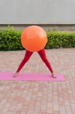 Spor hocası dışarıda pembe bir paspasın üzerinde turuncu bir fitness topuyla pilates yapıyor.