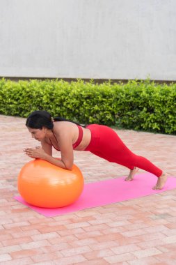 Genç bir kadın, asfalt bir alanda paspasın üzerinde spor topuyla pilates yapıyor.