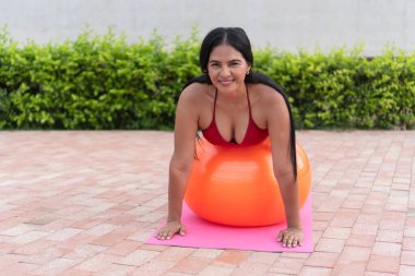 Latin bir kadın arka bahçede bir paspasın üzerinde spor topu kullanarak pilates yapıyor.