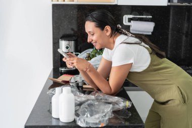 Ev hanımı geri dönüşüm için ev atıklarını ayırırken akıllı telefon kullanıyor ve gülümsüyor.