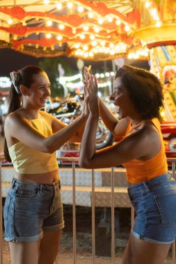 İki genç kadın el çırpıyor, lunaparkta eğleniyorlar.