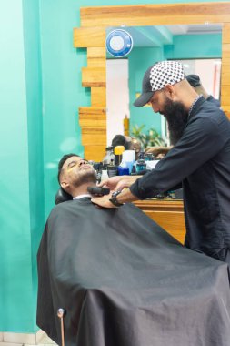 Berber, modaya uygun bir berber dükkanında müşterinin sakalını titizlikle buduyor, profesyonel bir tımar deneyimi sağlıyor.