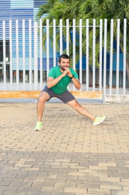 Spor kıyafeti giyen sakallı adam bacak esnetme hareketleri yapıyor, antrenmana hazırlanıyor.