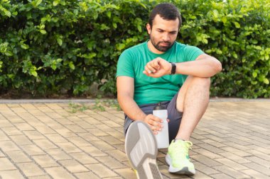 Yorgun sporcu antrenmandan sonra performansını kontrol ediyor, kaldırımda dinleniyor ve elinde su şişesi tutuyor.