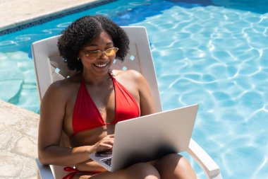 Kırmızı bikini ve güneş gözlüğü takıp havuz başında dizüstü bilgisayar kullanan serbest çalışan genç bir kadın iş hayatı dengesini gösteriyor.