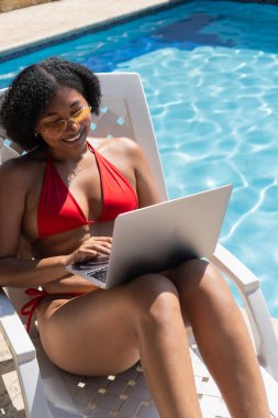 Genç bayan serbest yazar yaz tatilinde havuz kenarında iskemlede otururken dizüstü bilgisayarla çalışıyor.