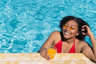 Kaygısız genç kadın güneşli bir günde berrak bir yüzme havuzunda dinlenirken ferahlatıcı bir portakal suyunun tadını çıkarıyor.