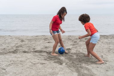 Sahilde futbol topuyla çıplak ayakla oynayan iki kız, deniz kenarında eğleniyorlar.