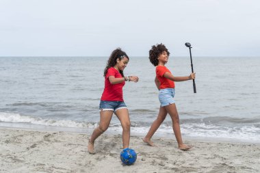 İki kız plajda selfie çubuğuyla futbol oynuyor ve video oynuyorlar.