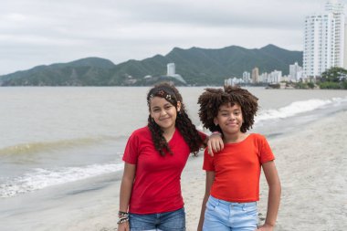 Arka planda deniz ve şehir olan kumlu bir plajda birlikte duran iki mutlu kız.