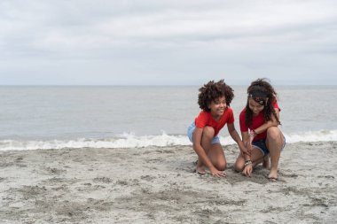 Çocukluğunda bulutlu bir sahilde kumla oynayan iki farklı genç kız.