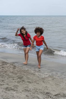 Okyanus kıyısında yürüyen afro saçlı iki gülümseyen kız.