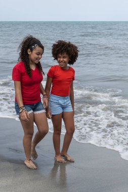 İki neşeli farklı kız okyanus dalgalarıyla kumlu bir sahilde yürüyüş yapıyor.